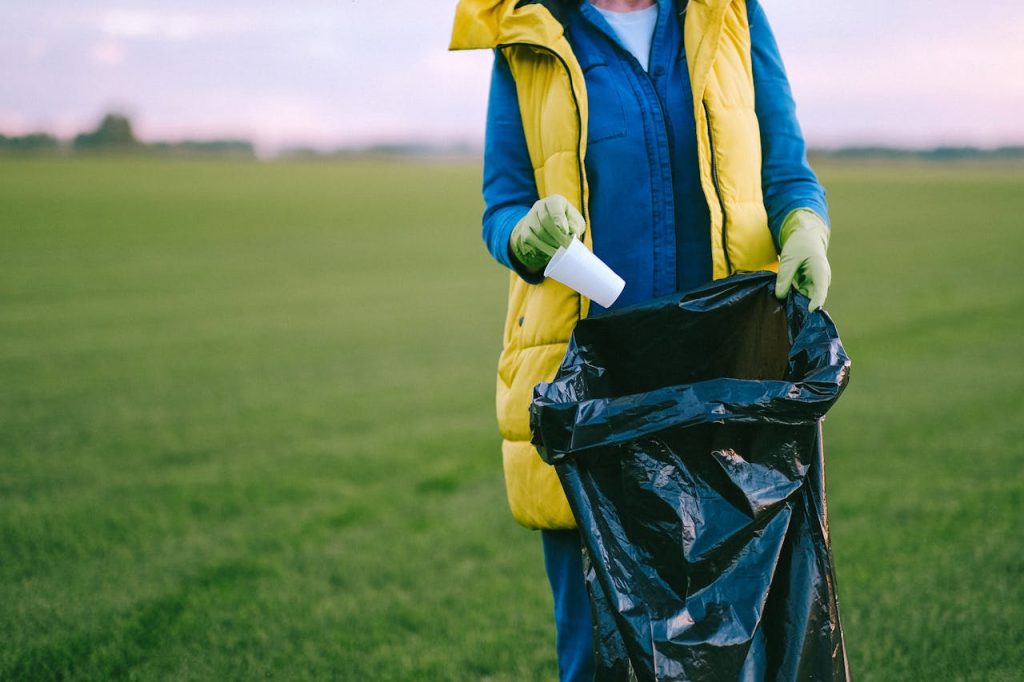 A volunteer cleans up a grass field by collecting trash into a black garbage bag.